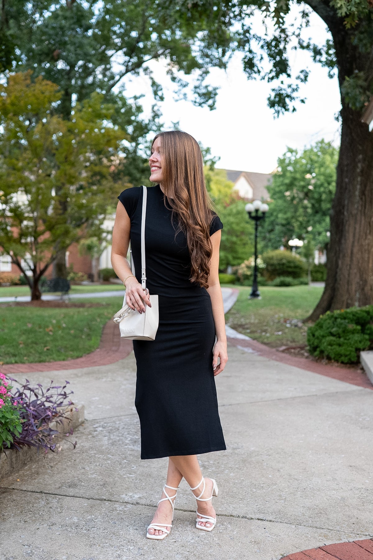 Woman wearing black bodycon dress with white strappy heels and a white purse, standing in a park