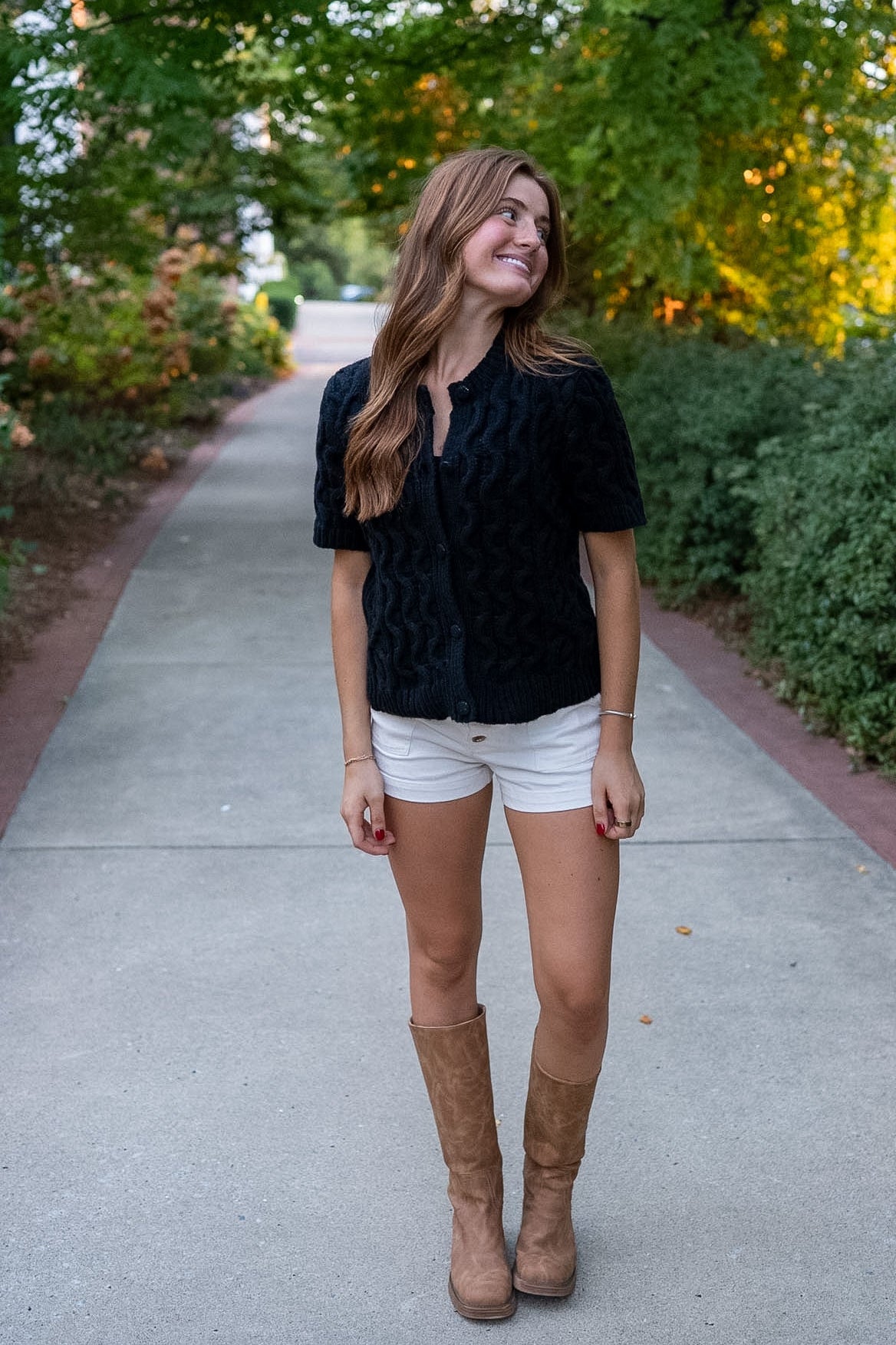 Woman wearing black short sleeve sweater with white shorts and tall boots standing on sidewalk surrounded by shrubs and trees.