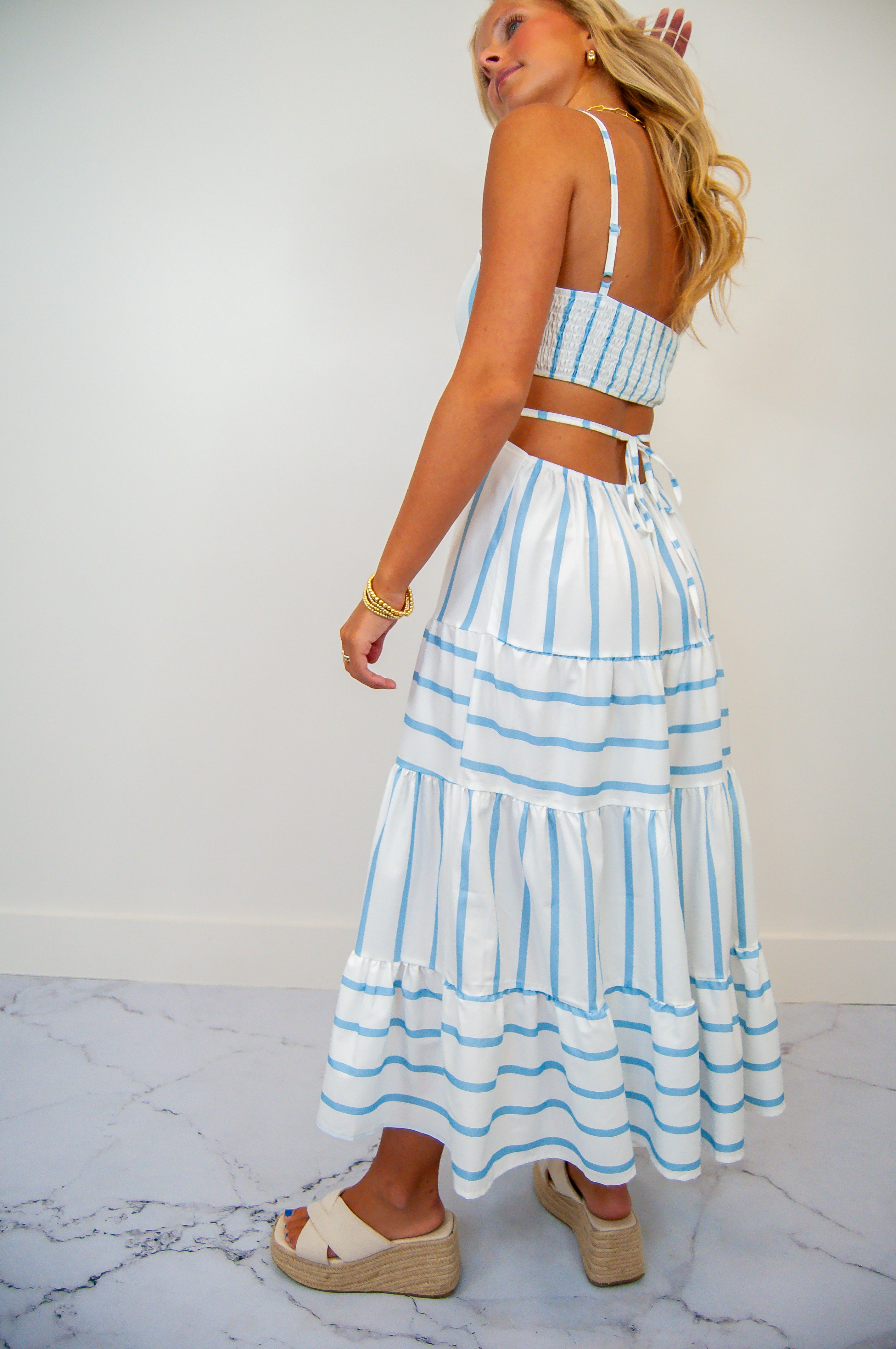 A woman wearing a backless sun dress with blue stripes and a cut-out back. The dress reaches mid-calf and she is wearing white sandals.