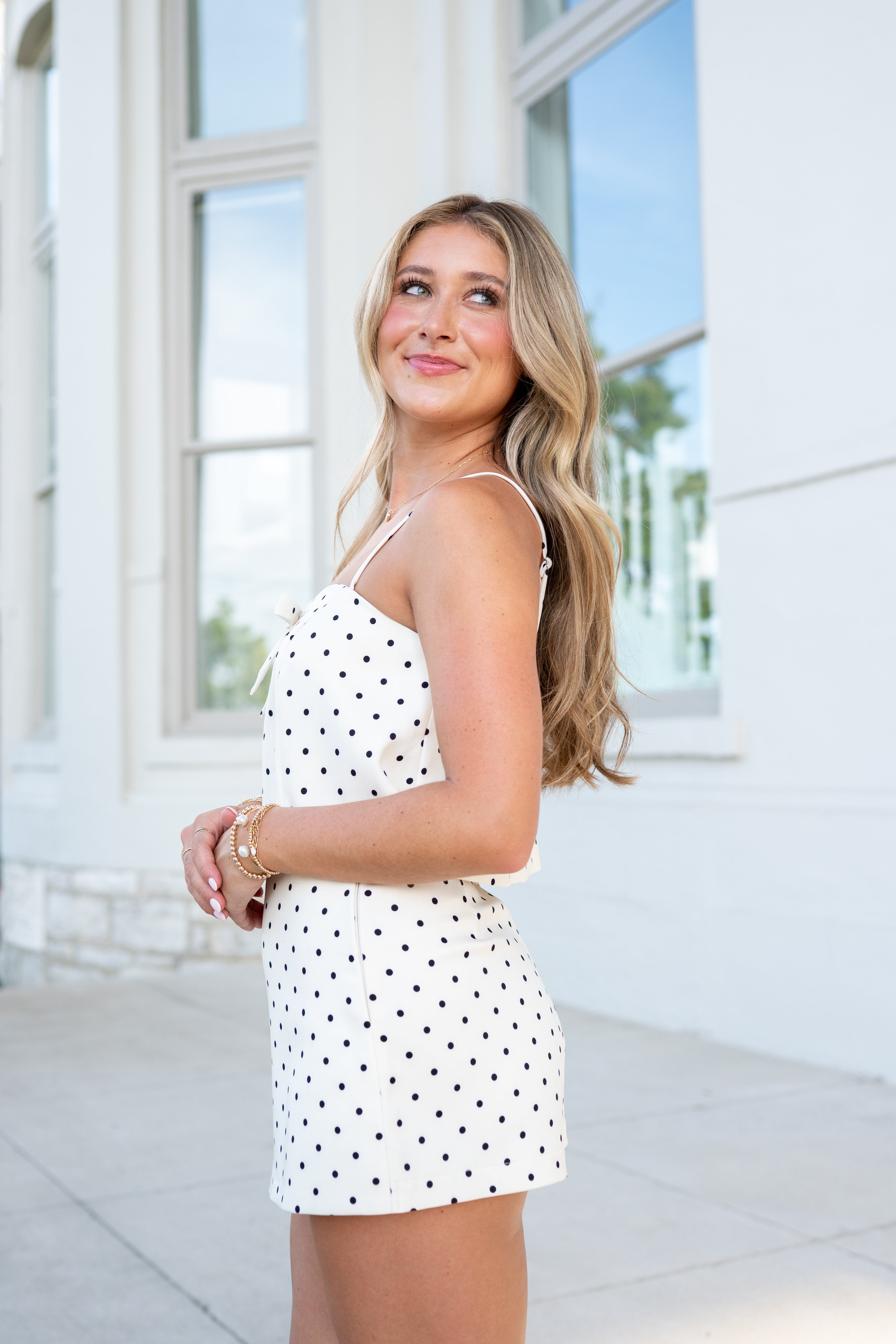 Woman wearing a white polka dot dress standing in a bright room with large windows.