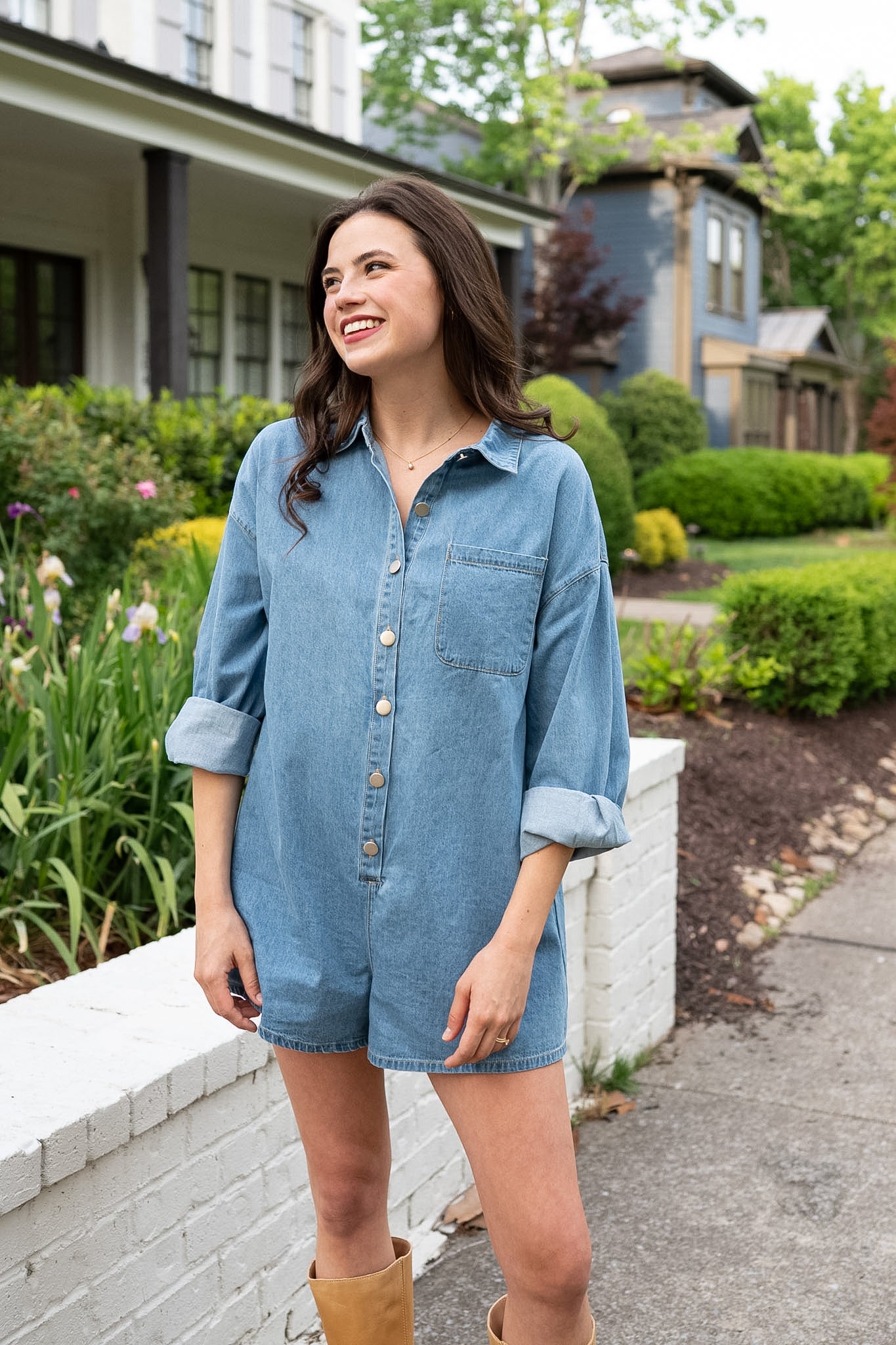 Woman wearing short denim romper and boots standing by white brick wall