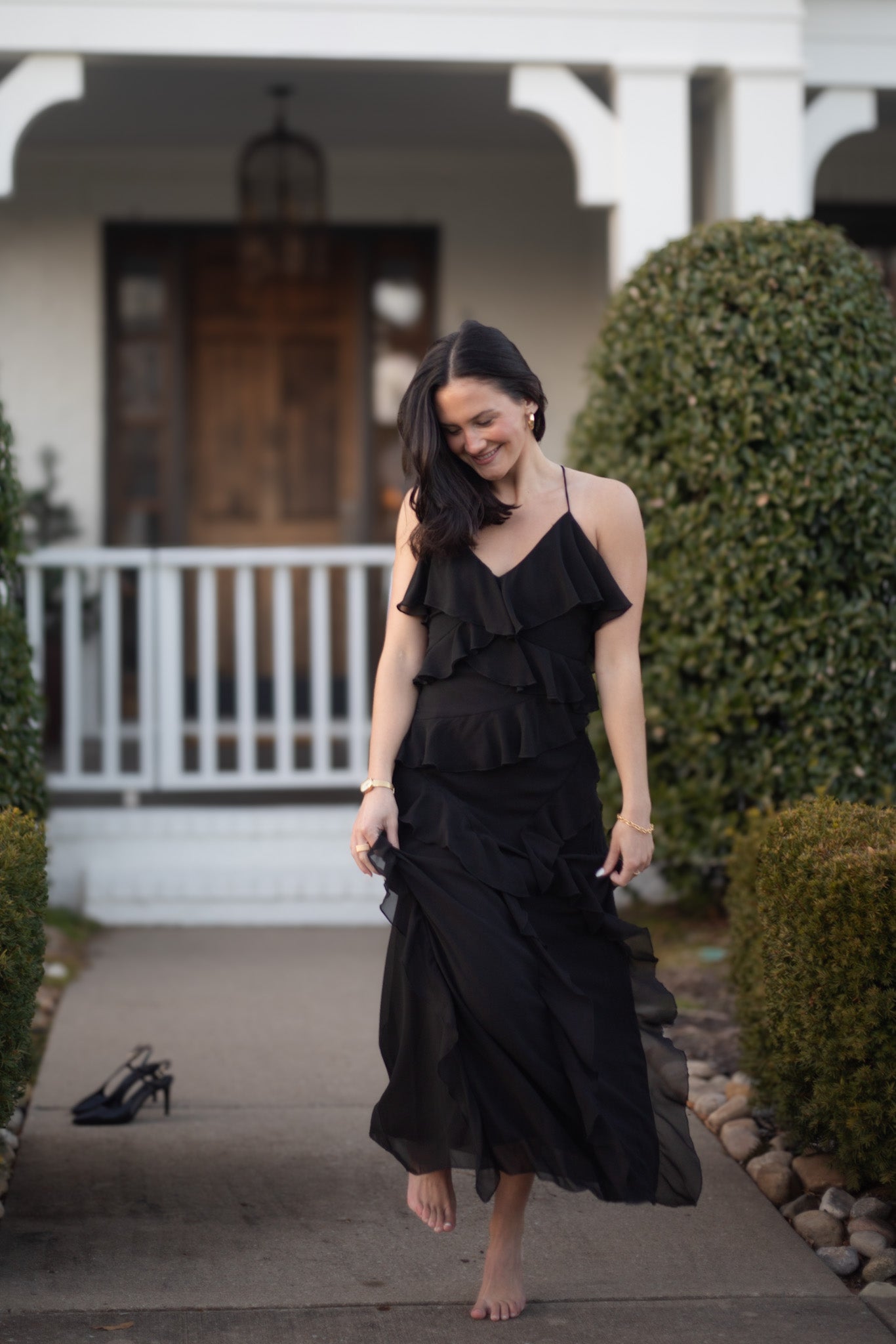 Woman in a black dress standing in front of a house with shoes nearby.