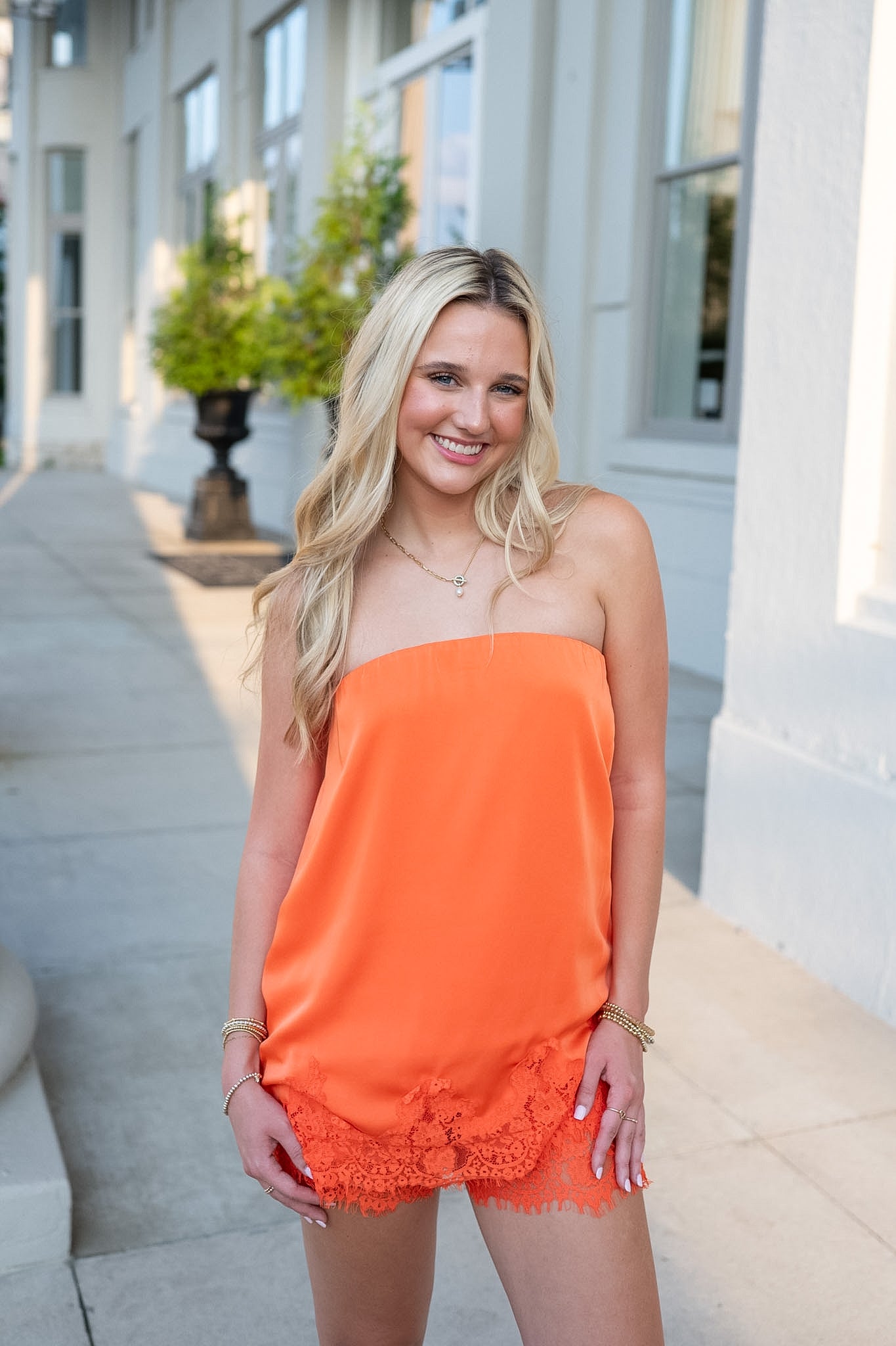 A smiling blonde woman wearing a bright orange strapless tube top with a lace hem, standing outdoors on a sidewalk in front of a white building.