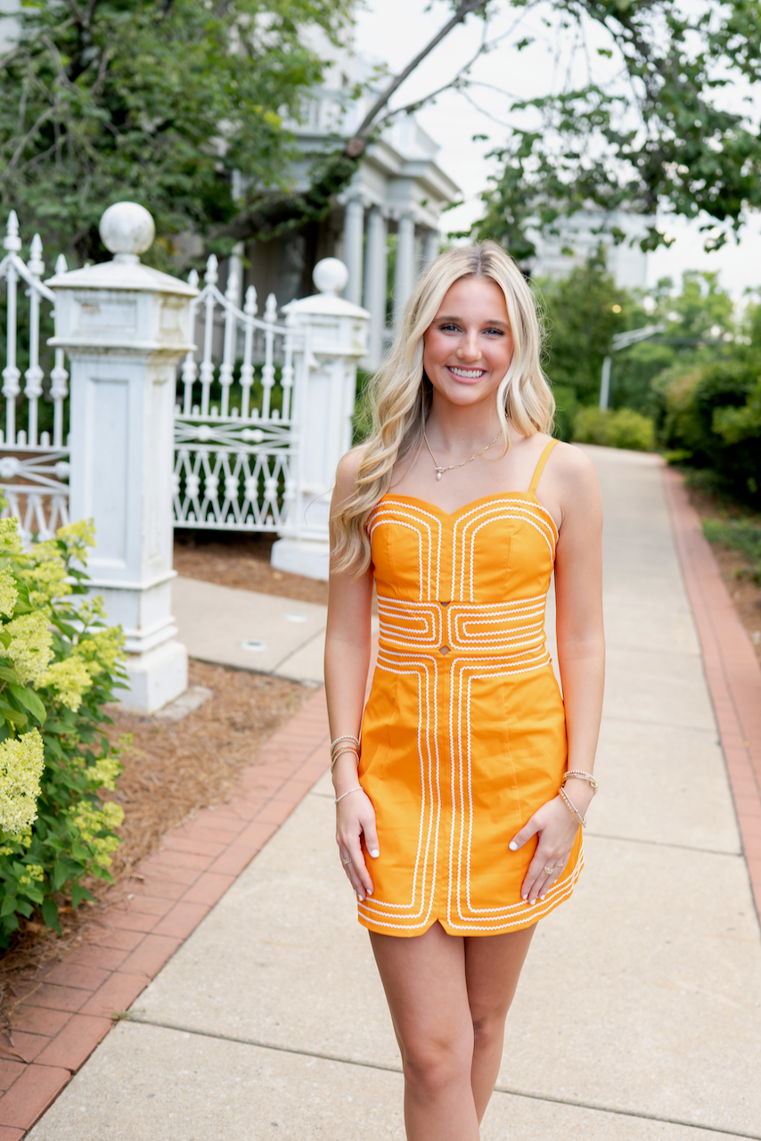 Woman in an orange dress standing on a sidewalk with greenery and a white fence in the background
