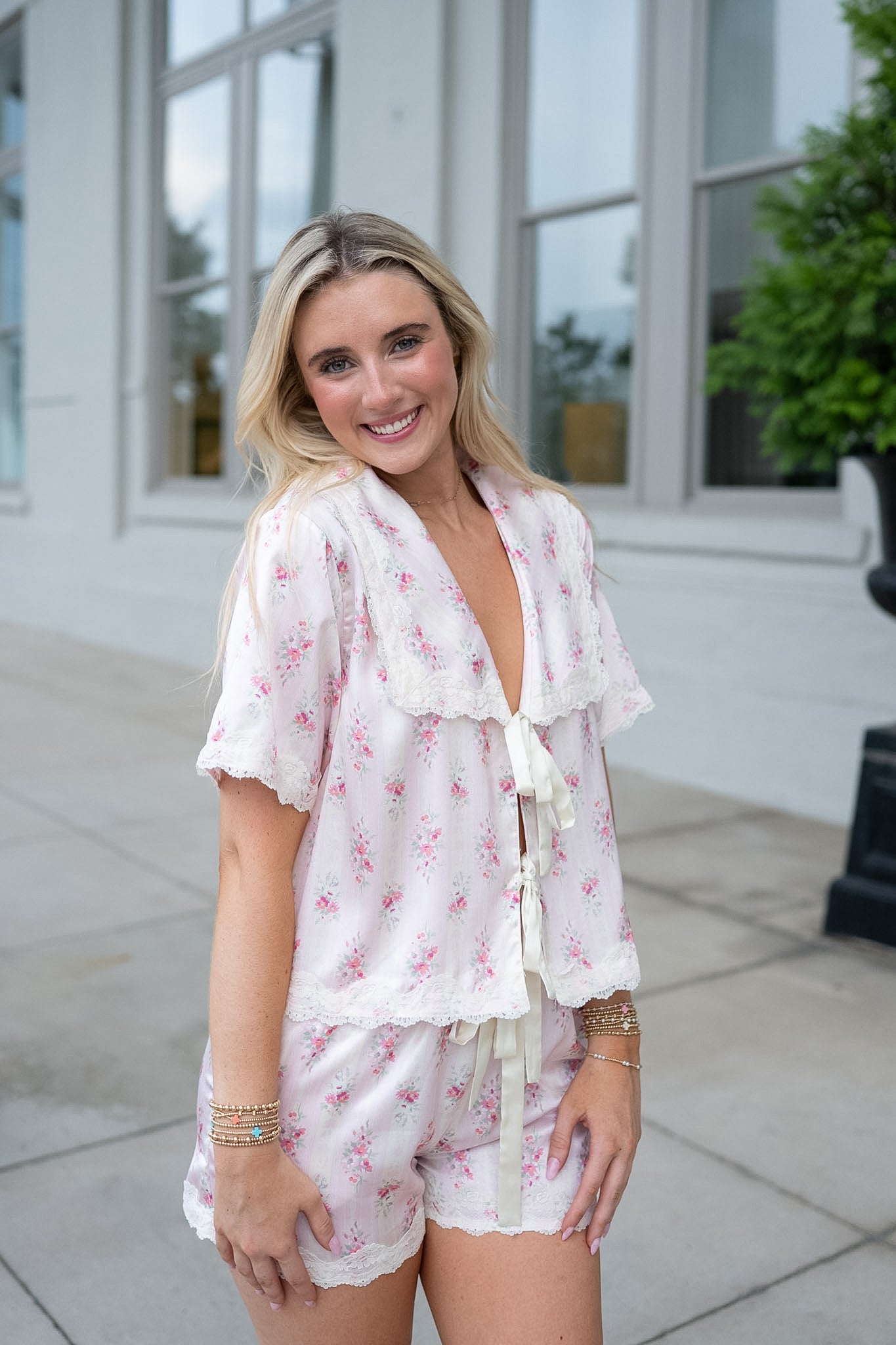 A woman modeling a two-piece pajama set featuring a white base with a pink floral print and ruffled lace trim, standing outdoors in front of a white building.