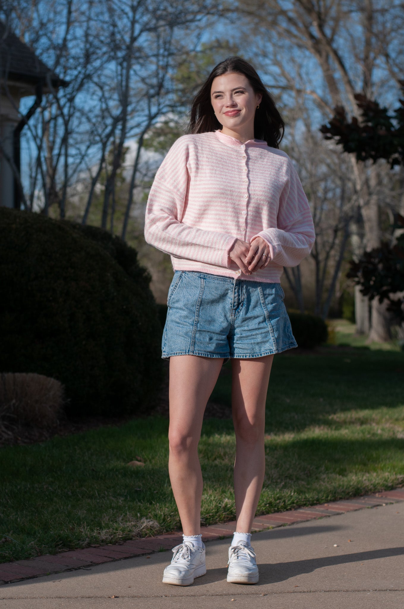 Woman wearing a pink sweater and denim shorts standing outdoors on a sunny day.