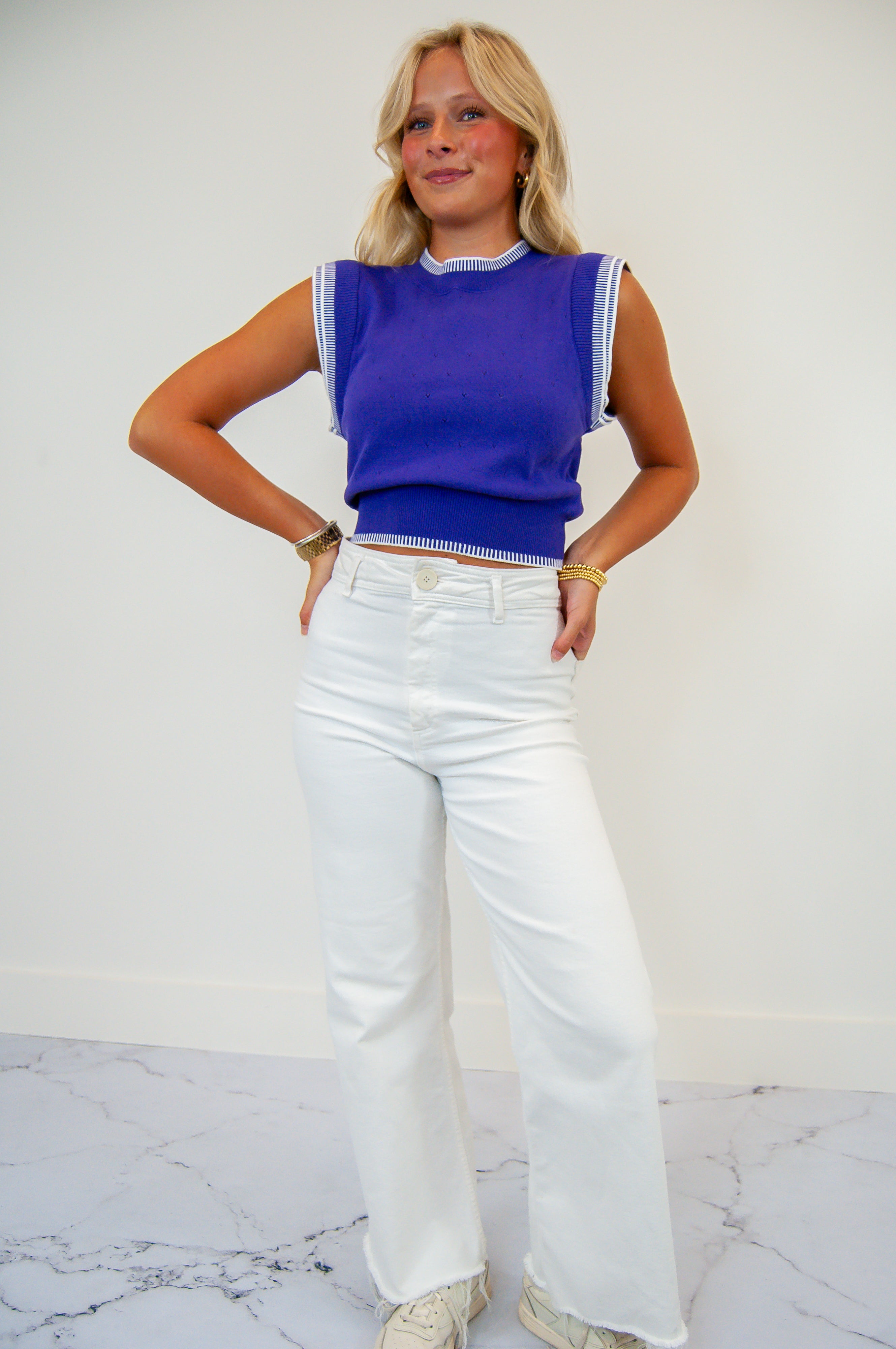 A person wearing a sleeveless purple top with white pants. The top has embroidered details on the shoulders.