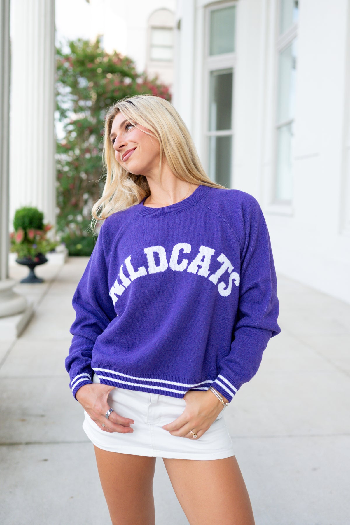 A woman wearing a purple crewneck sweatshirt with 'WILDCATS' printed in white arched letters across the chest, paired with a white mini skirt, standing outdoors on a sidewalk.