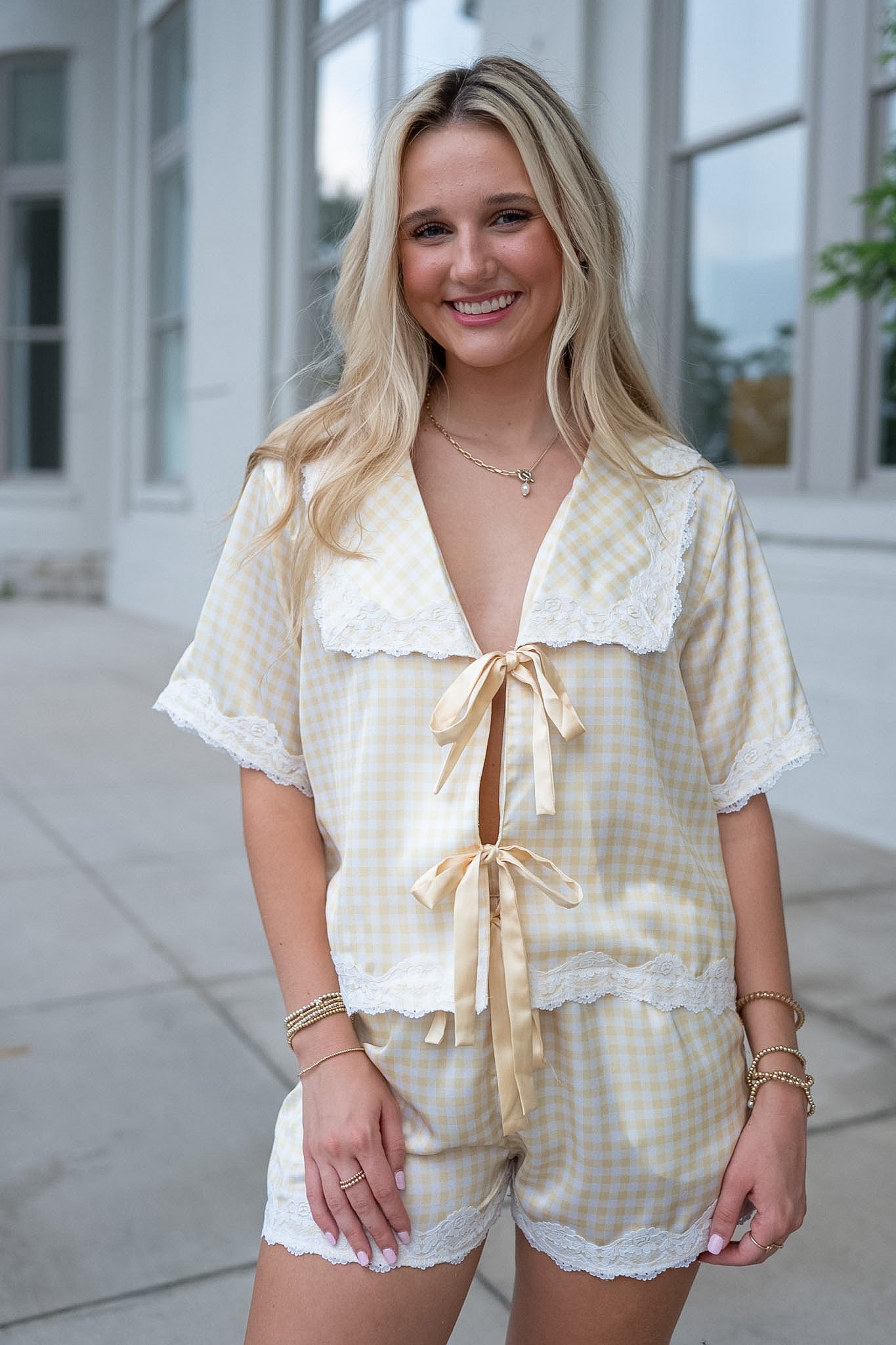 A smiling blonde woman wearing a light yellow and white gingham two-piece pajama set with white lace trim, featuring a short-sleeve top with a tie-front keyhole and matching shorts.