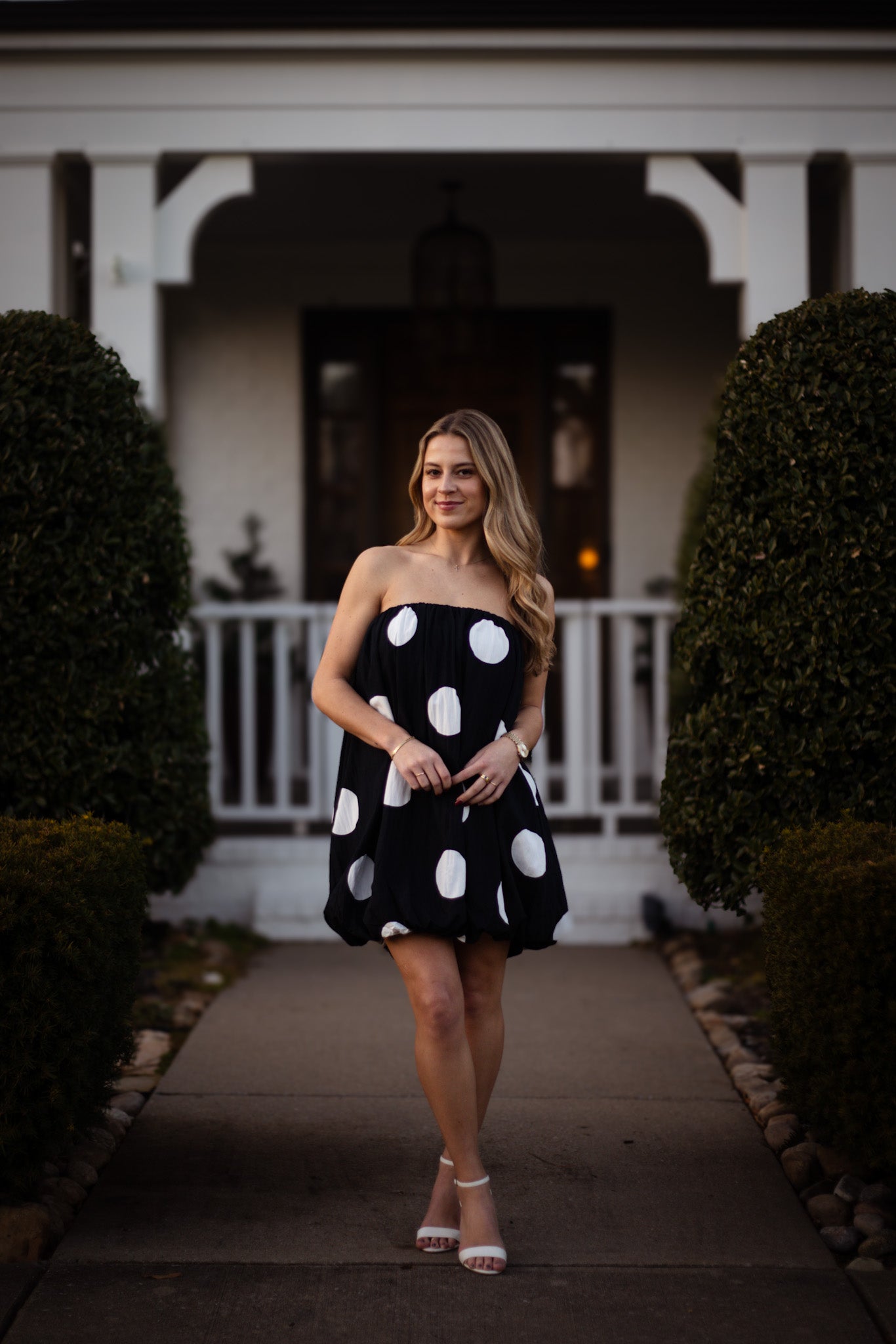 Woman in a black dress with white polka dots standing in front of a house.