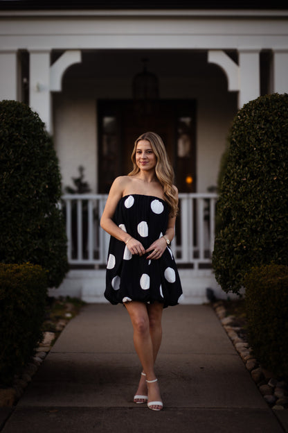 Woman in a black dress with white polka dots standing in front of a house.