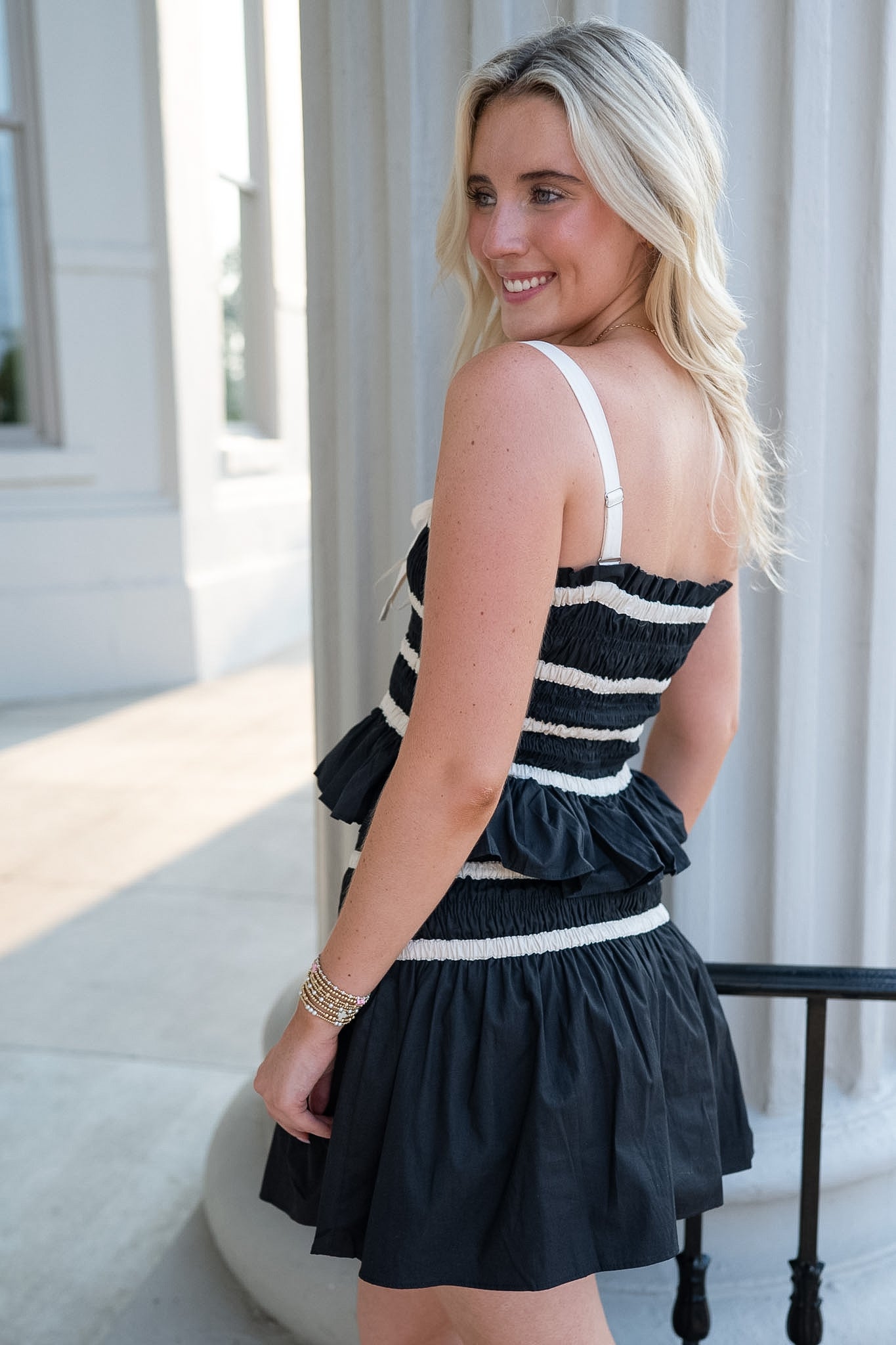 A woman wearing a black and white striped ruffled mini dress, photographed from behind in an outdoor patio setting with white columns.