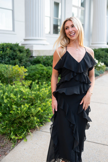 A woman wearing a black ruffled spaghetti-strap dress stands on a sidewalk in front of a white building with columns and green shrubs.