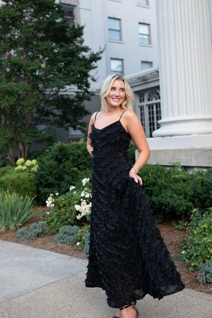 A woman wearing a black ruffled spaghetti-strap maxi dress stands outdoors in front of a white classical building with columns and green landscaping.