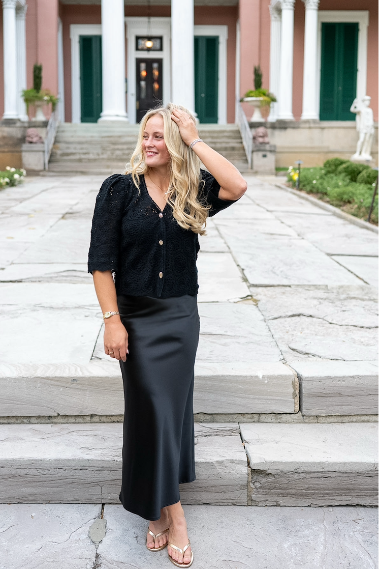 Woman wearing black satin midi skirt with black top standing on limestone patch in front of a pink and green building