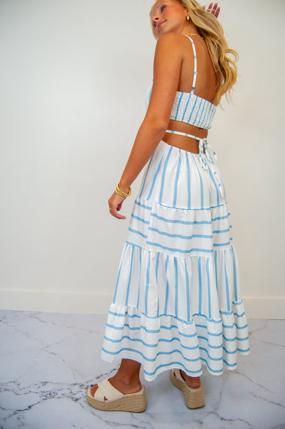 A woman wearing a backless sun dress with blue stripes and a cut-out back. The dress reaches mid-calf and she is wearing white sandals.