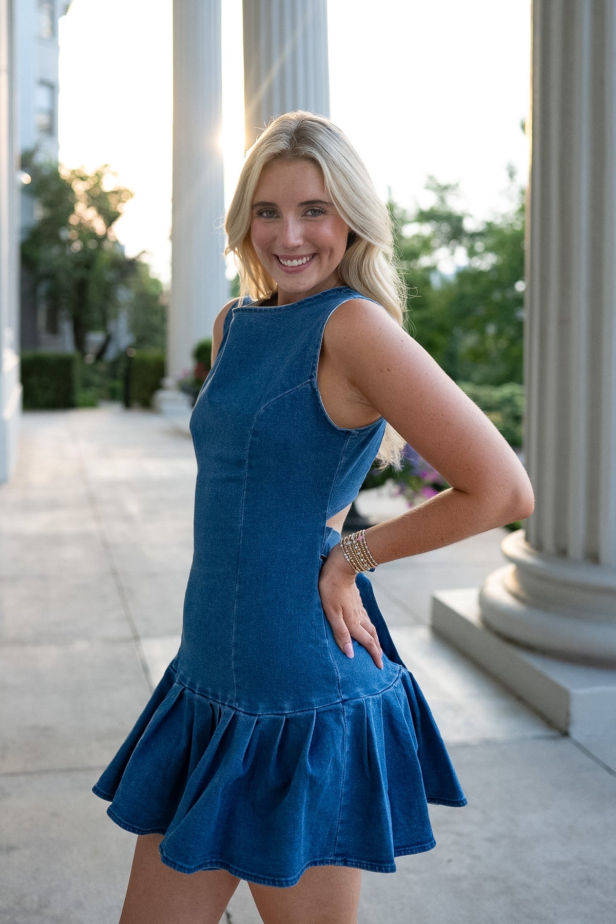 Girl wearing blue denim drop waist mini dress with wide pleats and tie back standing on a porch with white pillars.