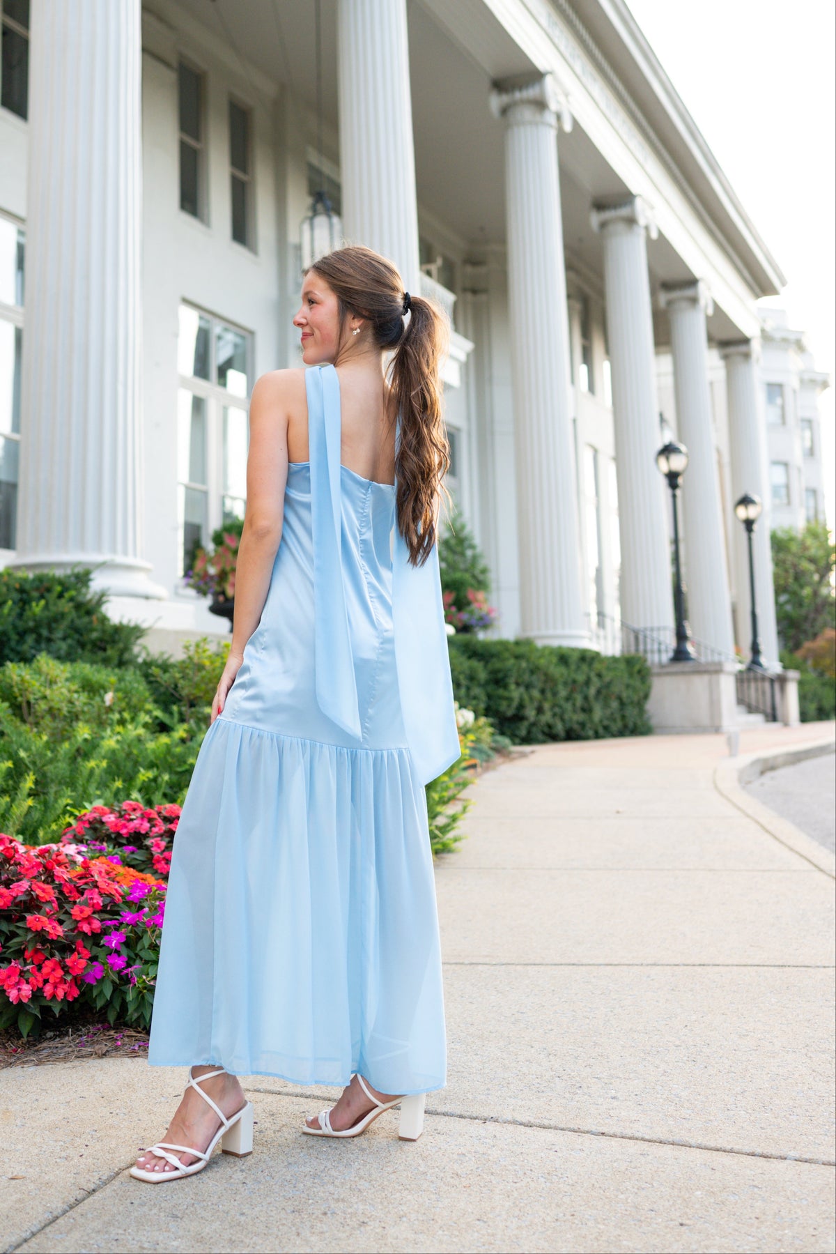 A woman wearing a light blue tiered maxi dress and white heeled sandals stands on a sidewalk in front of a white classical building with large columns.