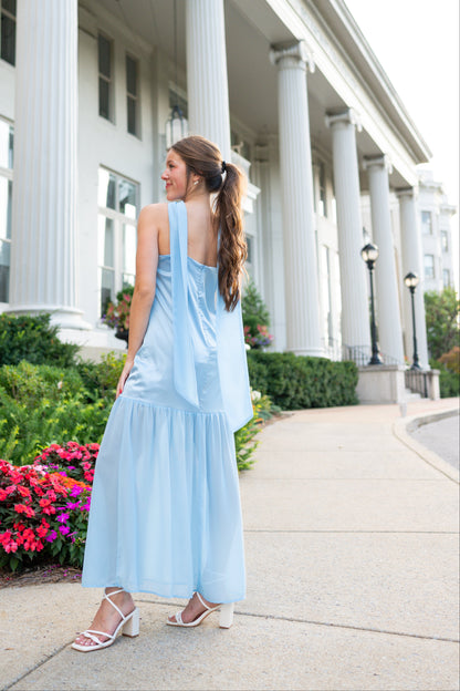 A woman wearing a light blue tiered maxi dress and white heeled sandals stands on a sidewalk in front of a white classical building with large columns.