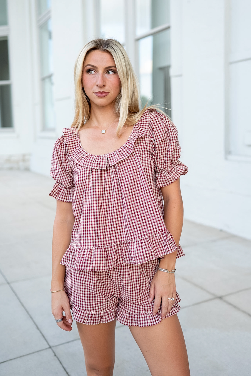 A blonde woman wearing a burgundy and cream gingham two-piece set consisting of a ruffled short-sleeve top and matching shorts, standing outdoors in front of a white building.