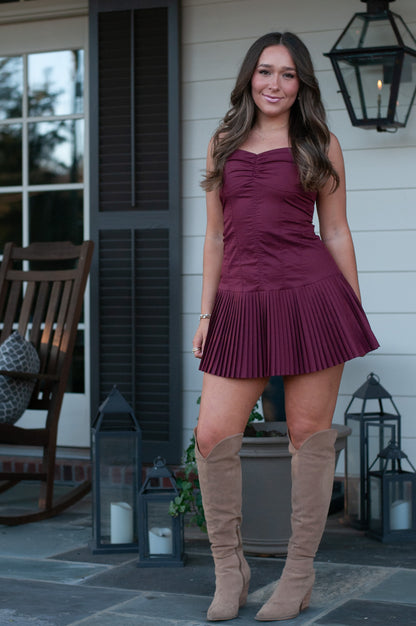 Woman wearing burgundy drop waist mini dress with knee high tan boots standing on a white porch