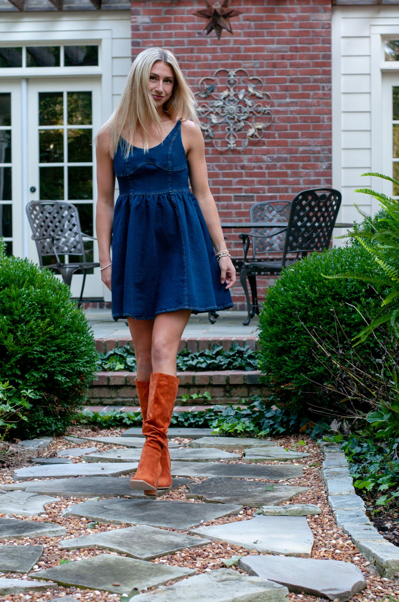 Woman in a blue denim dress and orange  brown boots standing on a stone path in front of a brick building.