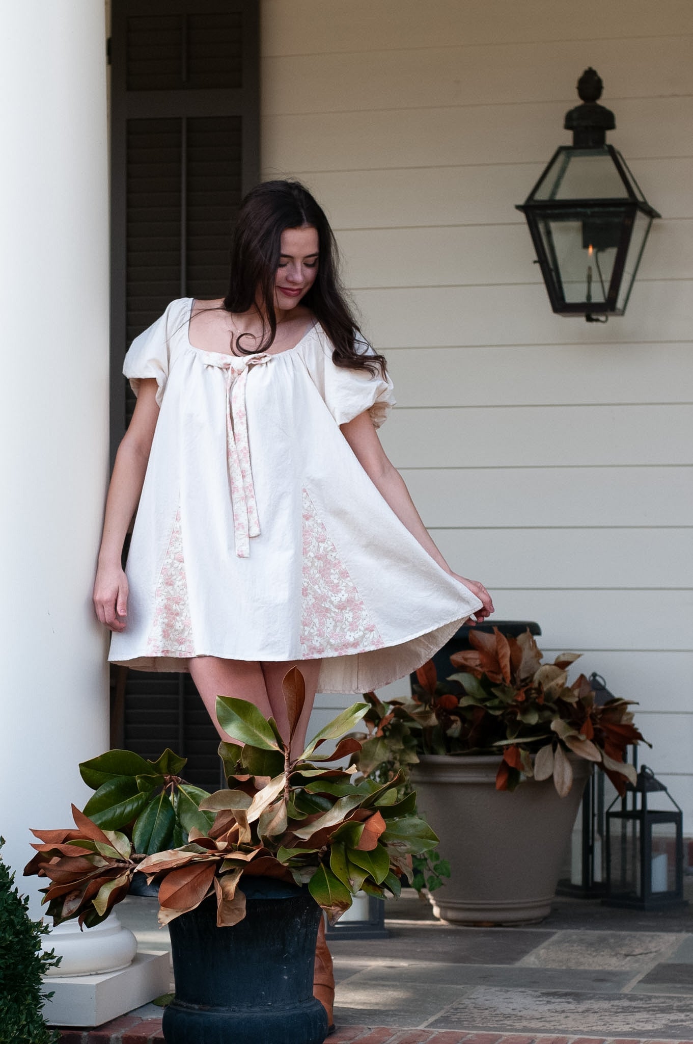 Woman in a white dress standing on a porch with decorative plants and lanterns.