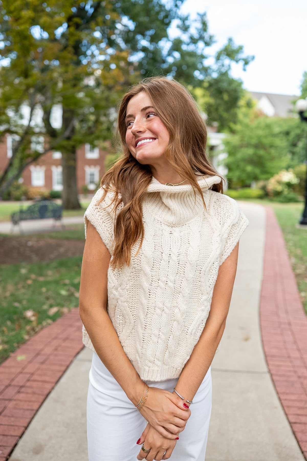 Woman wearing ivory sleeveless sweater standing on sidewalk by trees.