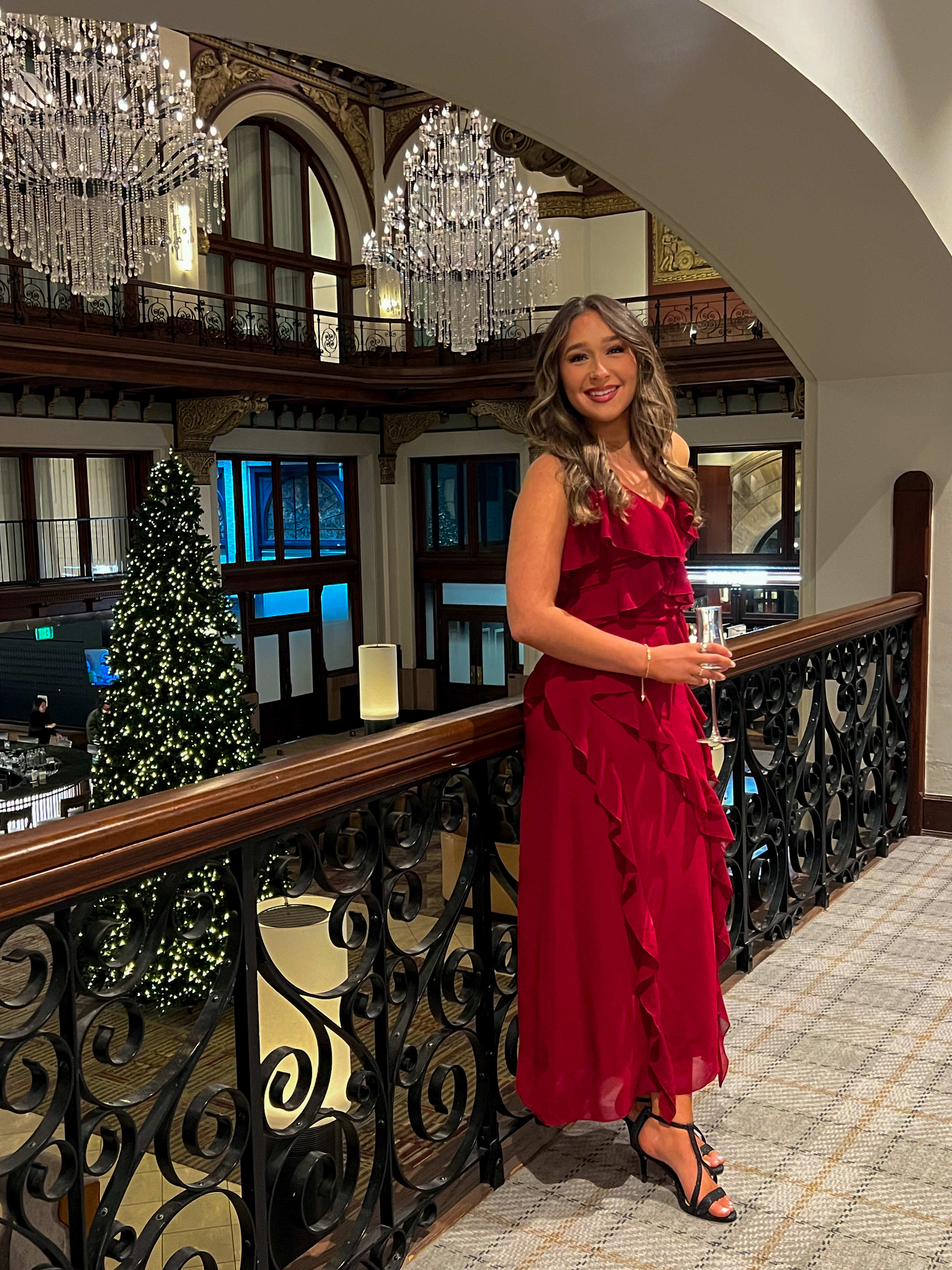 Woman in a red dress standing in an elegant interior setting with chandeliers and a Christmas tree.