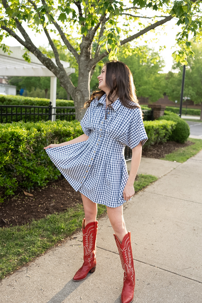 Woman in a checkered dress and red cowboy boots standing on a sidewalk with greenery in the background