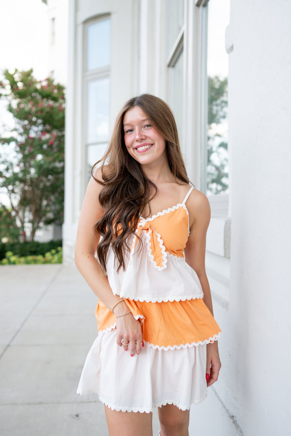 A young woman smiling and posing outdoors in a two-piece orange and white ruffled outfit consisting of a spaghetti-strap top and a matching mini skirt.