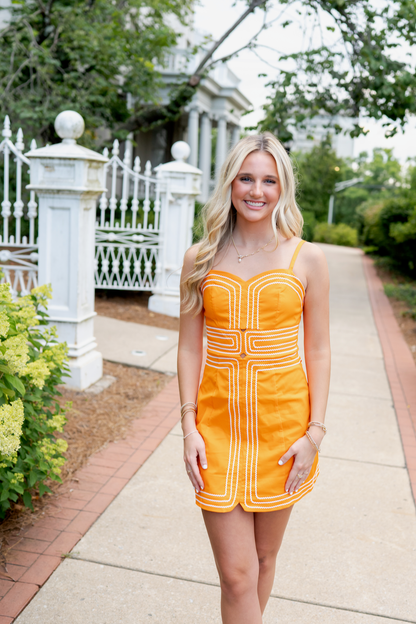 Woman in an orange dress standing on a sidewalk with greenery and a white fence in the background