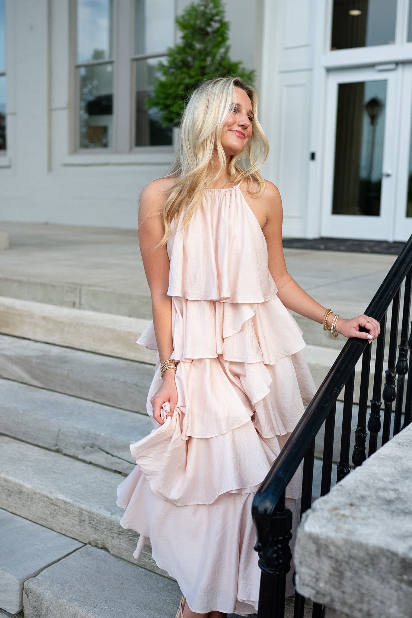 A woman wearing a pale blush pink halter-neck ruffled dress stands on outdoor stone steps next to a black metal railing in front of a white building.