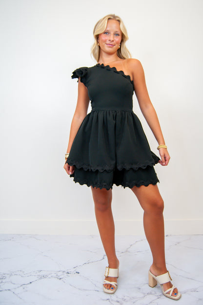 A woman wearing a one shoulder scallop edge mini dress with matching ric rac trim in black color.