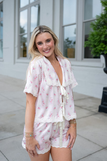 A woman modeling a two-piece pajama set featuring a white base with a pink floral print and ruffled lace trim, standing outdoors in front of a white building.