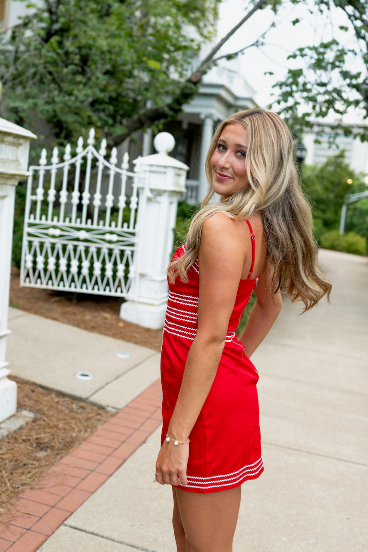 Woman in a red dress standing on a sidewalk with a white fence and trees in the background