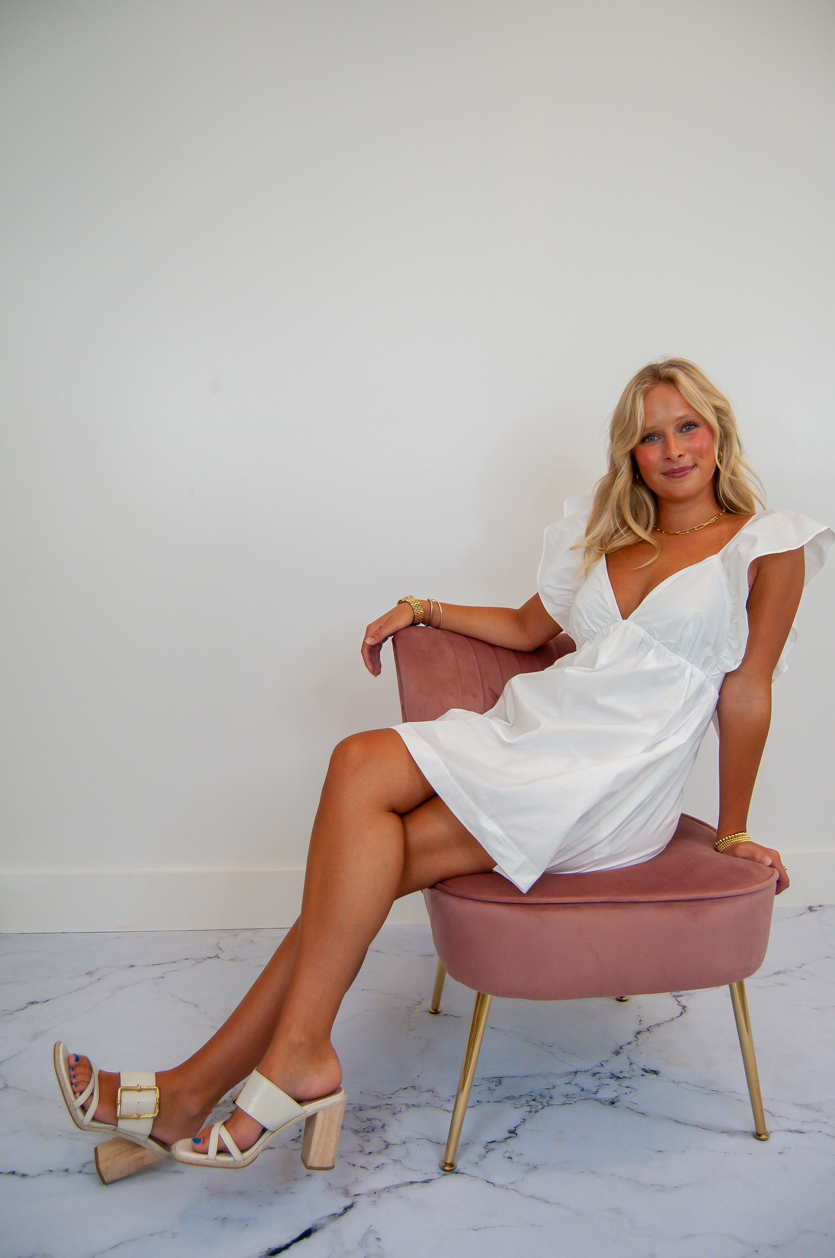 A woman seated on a pink chair wearing a white mini dress with a ruffle design and tie back detail.