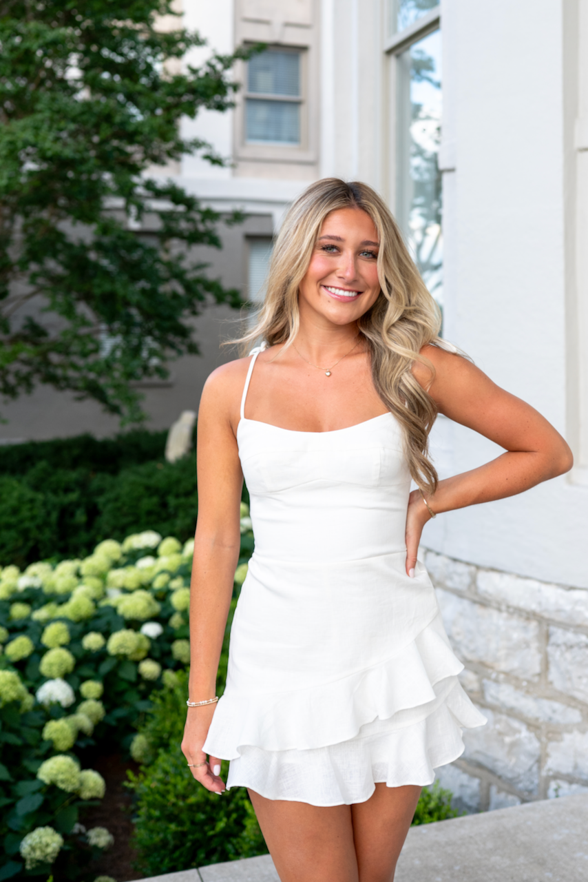 Woman in a white dress standing outdoors with greenery and a building in the background