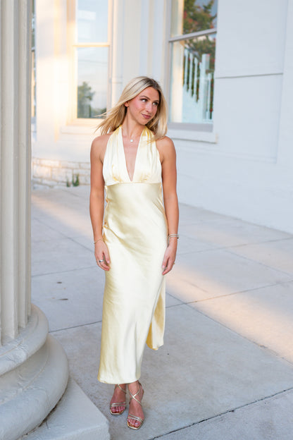 A woman wearing a pale yellow satin halter midi dress with a front slit, standing on a sunlit stone walkway next to a white building with columns.