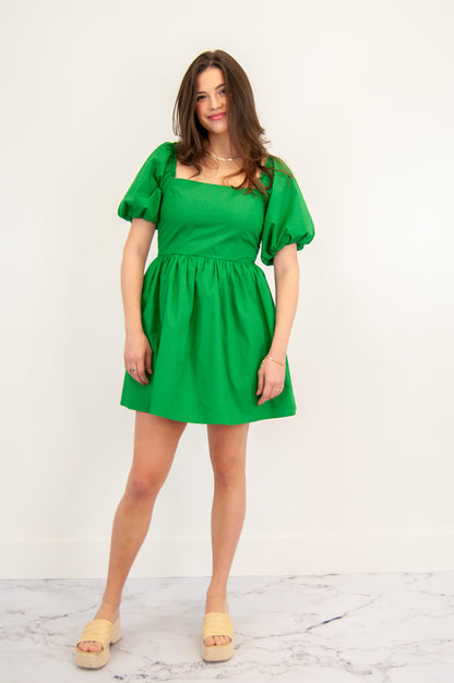 A woman wearing a short-sleeved square neck green dress with puff sleeves and a fitted bodice. She is standing against a white background.