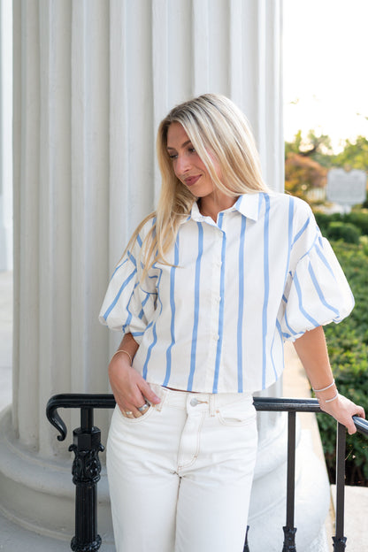 A blonde woman wearing a white and light blue vertically striped puff-sleeve blouse and white pants, standing on a balcony with white columns and greenery in the background.