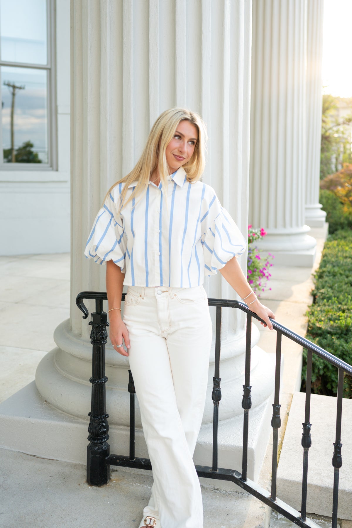 A woman stands on outdoor steps in front of large white columns, wearing a white puff-sleeve button-up shirt with thin blue vertical stripes and white wide-leg pants.